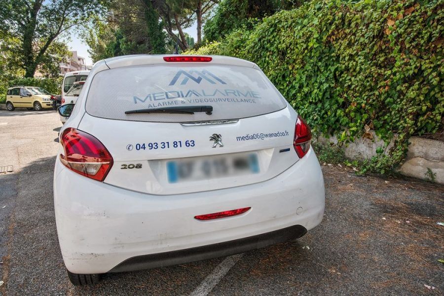 Arrière d'une Peugeot 350 blanche avec inscription sur la lunette arrière, garée en bordure de route bordée de haies.