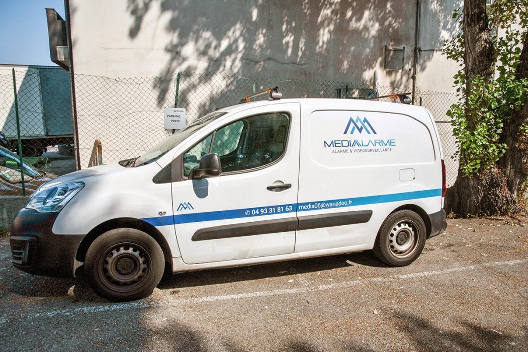 Fourgonnette de service blanche garée contre un mur, avec logo et texte bleus sur le côté.