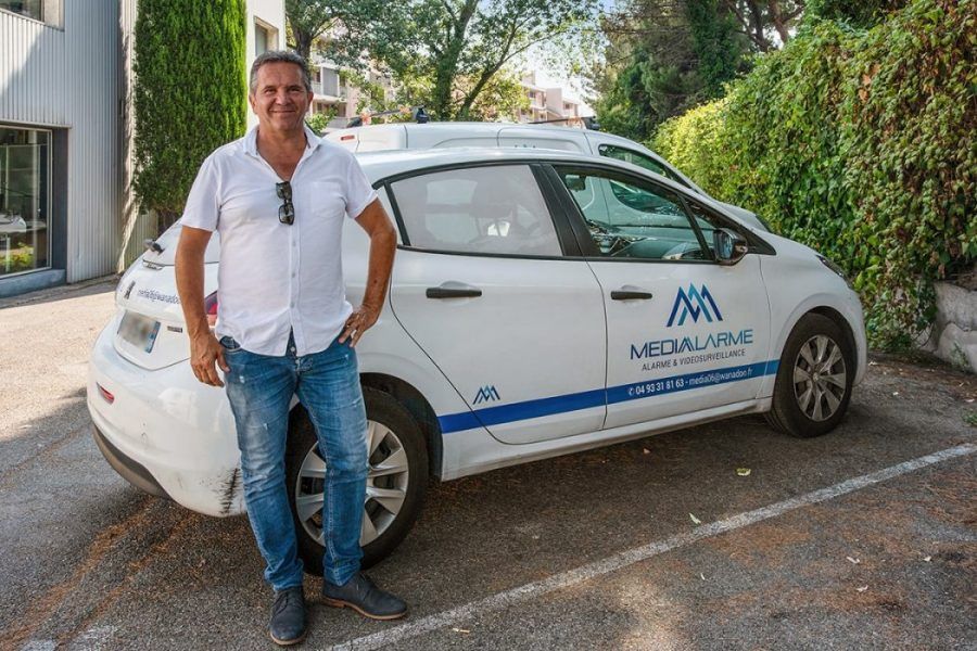Un homme se tient debout à côté d'une voiture blanche avec un logo et une bande bleue dans une rue résidentielle.