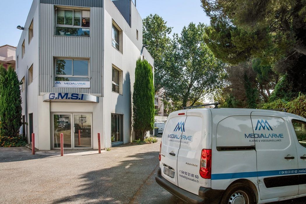 Immeuble de bureaux blanc avec enseignes bleues et camionnette blanche de l'entreprise garée devant, dans une rue ensoleillée.