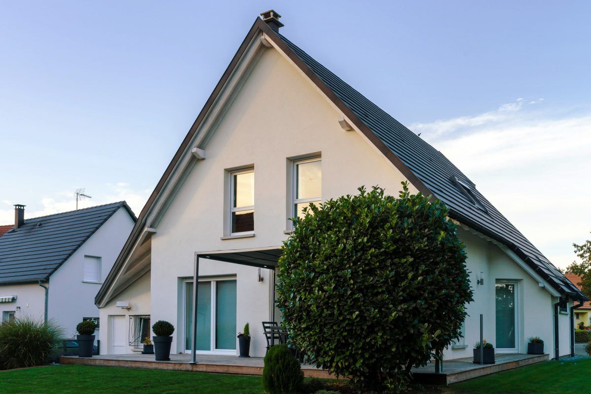 Maison blanche moderne avec un toit foncé, de grandes fenêtres et un arbre taillé devant.