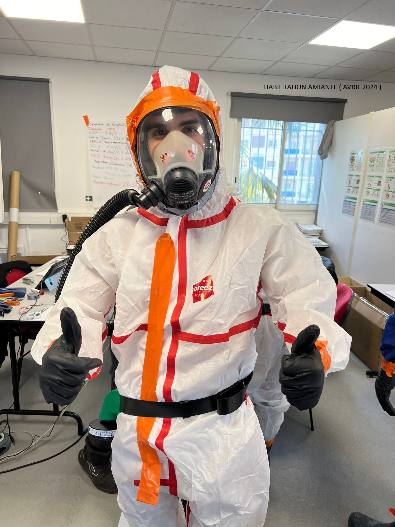 Une personne vêtue d'une combinaison de protection blanche et portant un masque respiratoire se tient dans une pièce ressemblant à une salle de classe.