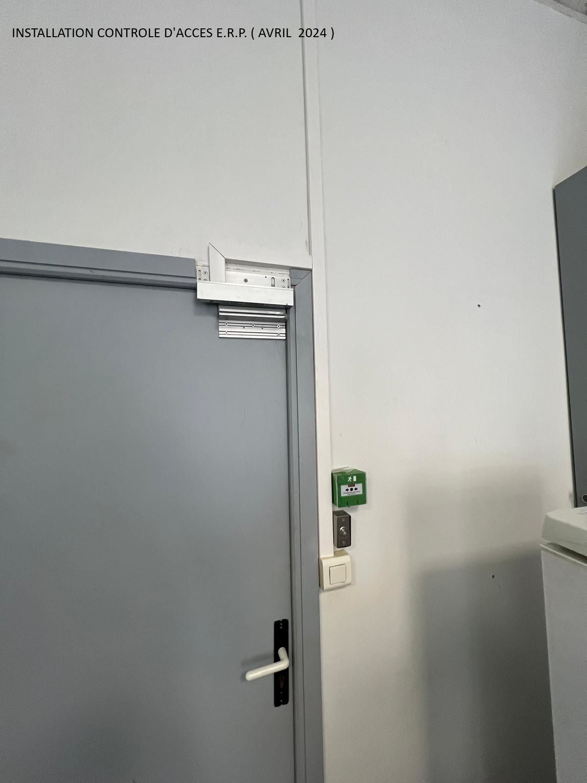 Porte grise dans un couloir banal, avec un bouton mural vert à côté.