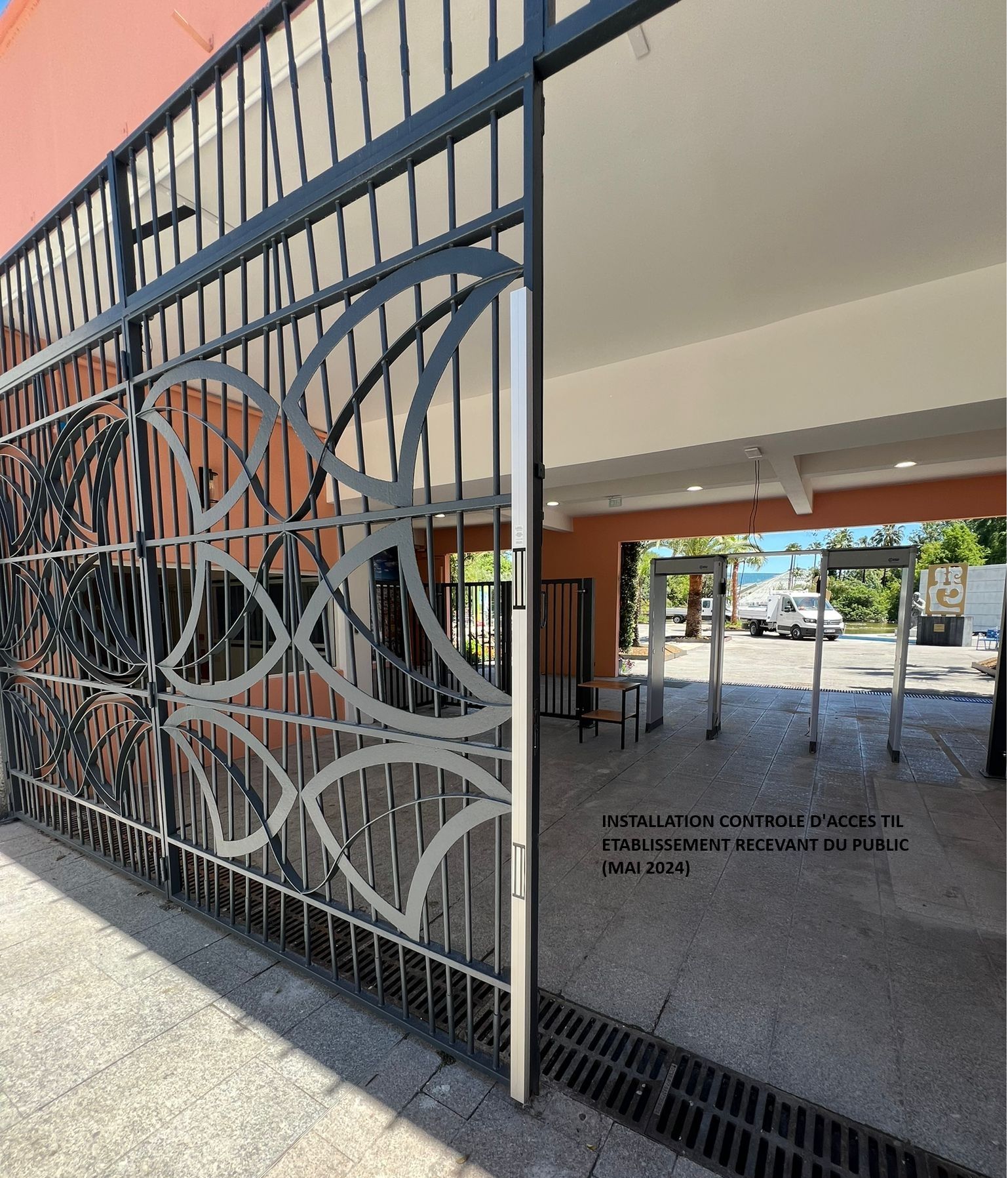 Portail ouvert donnant sur un parking avec des barres décoratives en métal et une cour ensoleillée au-delà.