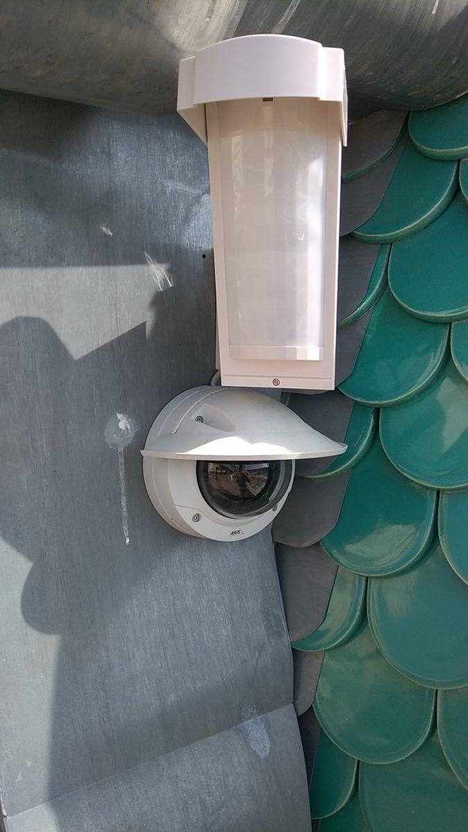 Caméra de sécurité extérieure fixée au mur, sous un éclairage cylindrique blanc sur un mur recouvert de bardeaux gris et verts.