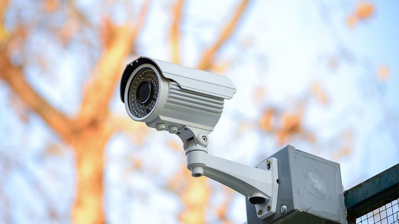 Caméra de sécurité extérieure fixée sur un poteau, pointée vers le ciel près d'arbres aux teintes automnales orangées