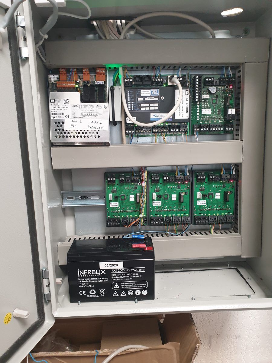 Panneau de commande électrique ouvert avec des circuits imprimés verts, des câbles et une batterie noire à l'intérieur d'une armoire métallique.