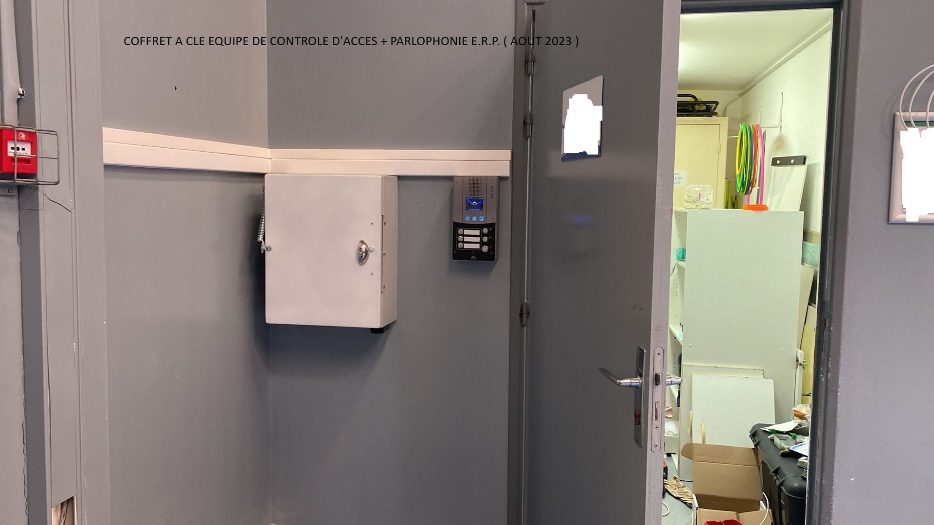 Mur gris avec boîtiers et clavier à code fixés au mur, à côté d'une porte ouverte donnant sur une buanderie éclairée.