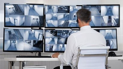 Une personne surveille les flux vidéo de plusieurs caméras de sécurité sur de grands écrans dans une salle de contrôle.