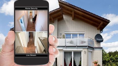 Main tenant un smartphone montrant les images d'une caméra de sécurité dans un couloir, à côté d'une maison avec une antenne parabolique.