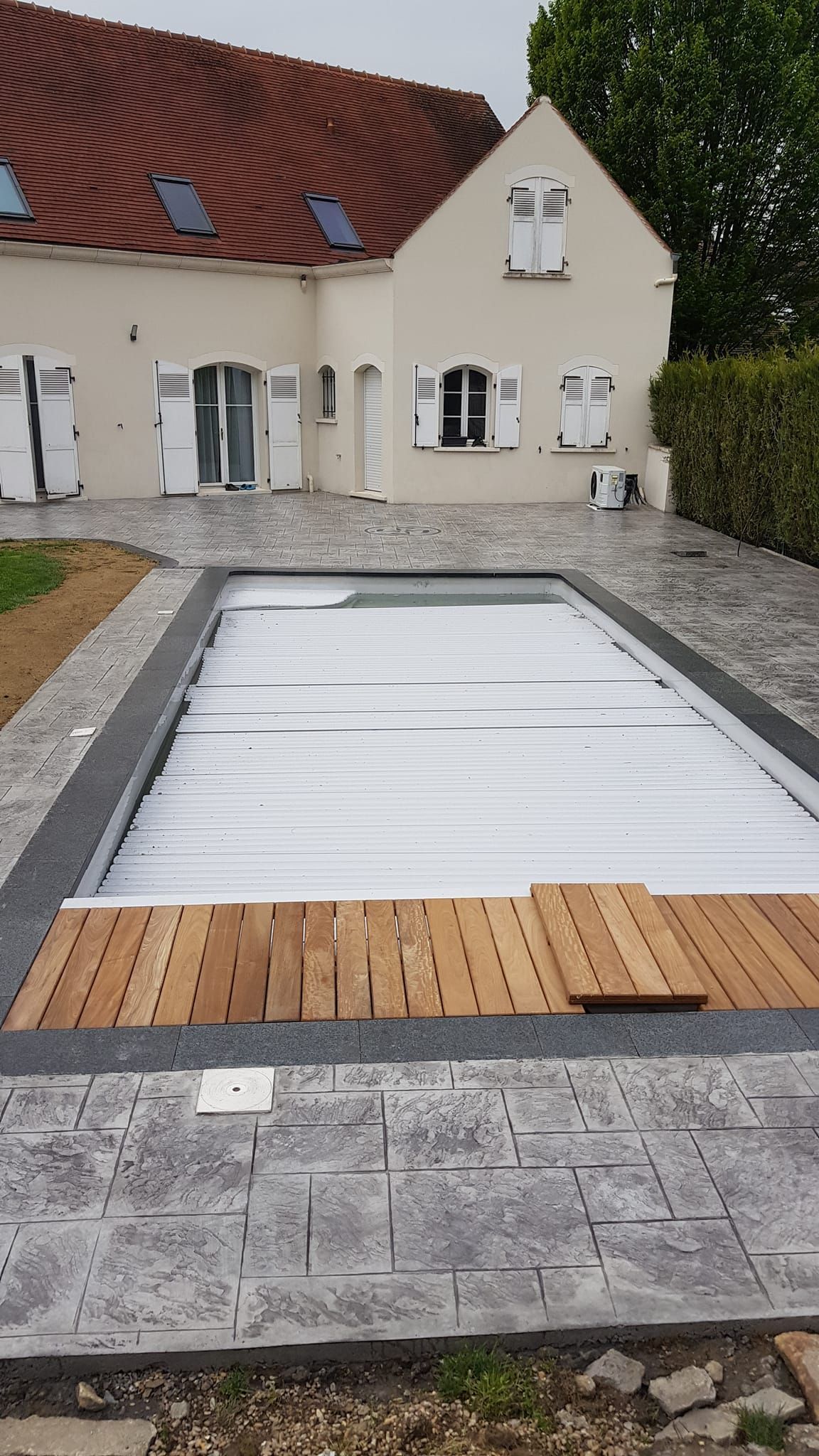 Une maison beige aux volets blancs avec une terrasse en béton imprimé de couleur grise.