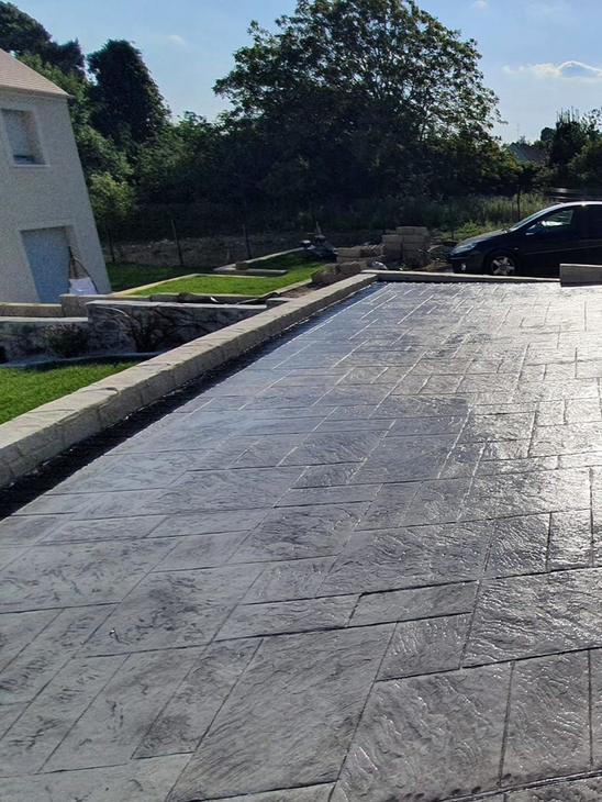 Une terrasse en béton imprimé grise, attenante à une maison beige, une voiture noire est garée devant.