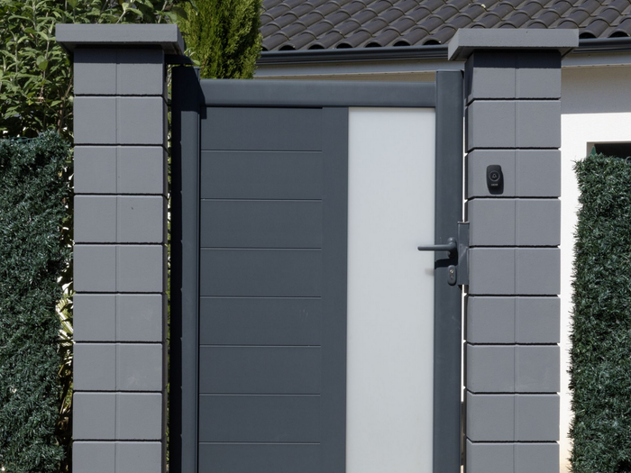 Vers la page Maçonnerie. Un portillon gris anthracite entre deux piliers en pierre couleur grise.