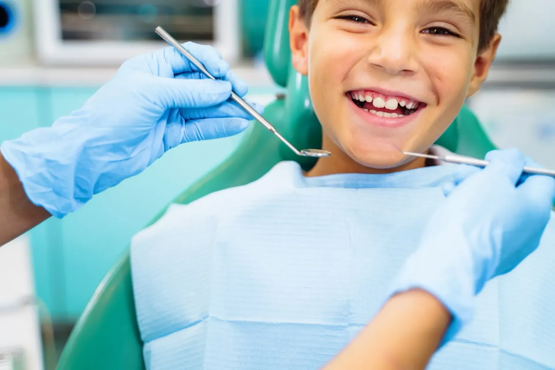 Niño sentado en el sillón del dentista sonríe durante el examen; se ven los guantes azules y las herramientas.