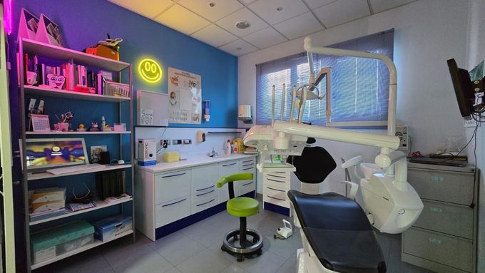 Consultorio dental con paredes azules, sillón dental y estanterías. Se ve un letrero de neón.