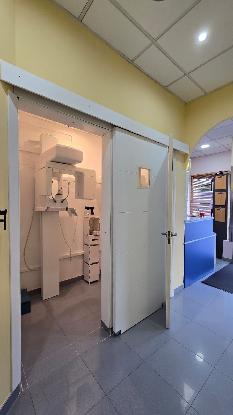 Pasillo de una consulta médica con suelo de baldosas grises, paredes amarillas y puerta abierta al equipo.