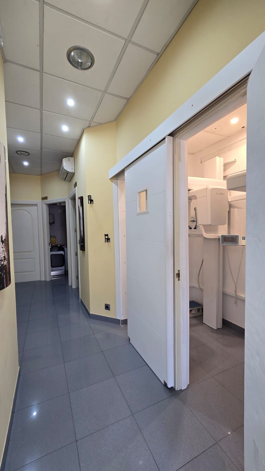 Pasillo de una consulta médica con suelo de baldosas grises, paredes amarillas y puerta abierta al equipo.