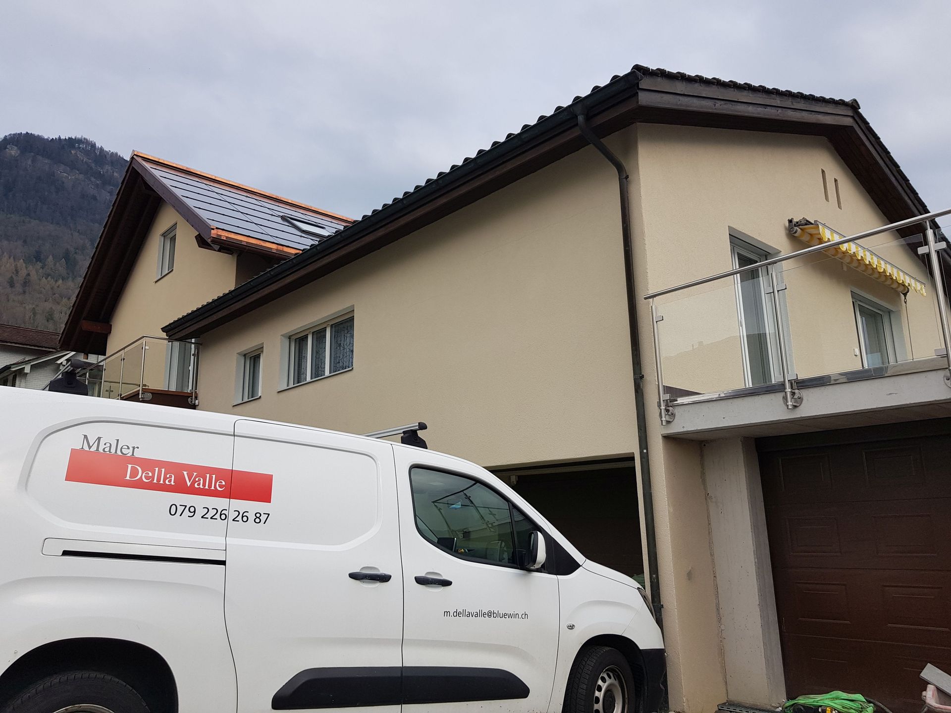 Weißer Lieferwagen, geparkt vor einem zweistöckigen beigen Haus mit braunem Dach und Balkon, vor einem bewölkten Himmel.