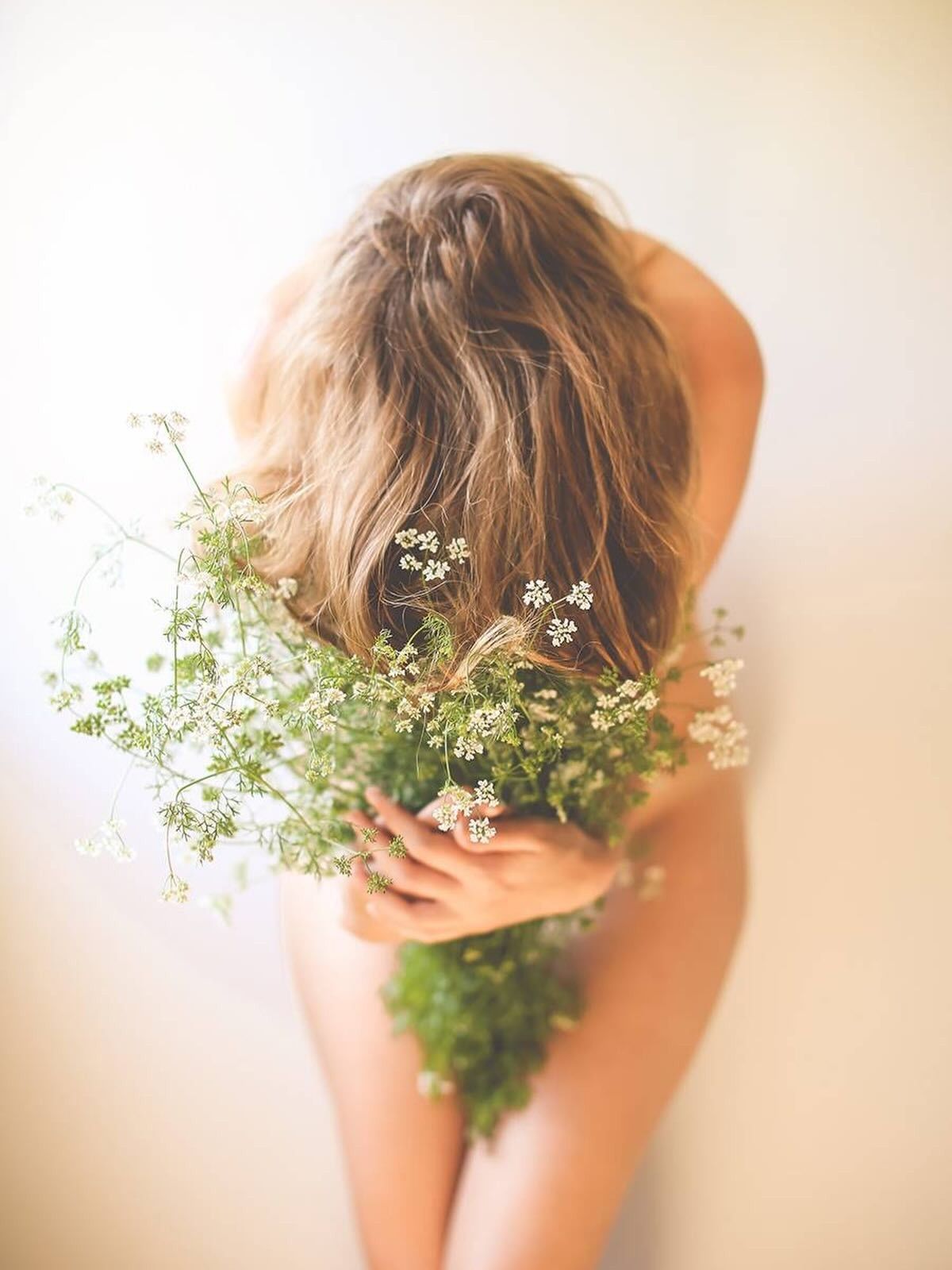 Persona con cabello largo posando desnuda, sosteniendo un ramo de flores blancas sobre un fondo brillante.