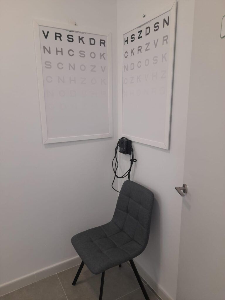 Sala de examen ocular con optogramas en la pared, una silla gris y un foróptero en la pared.
