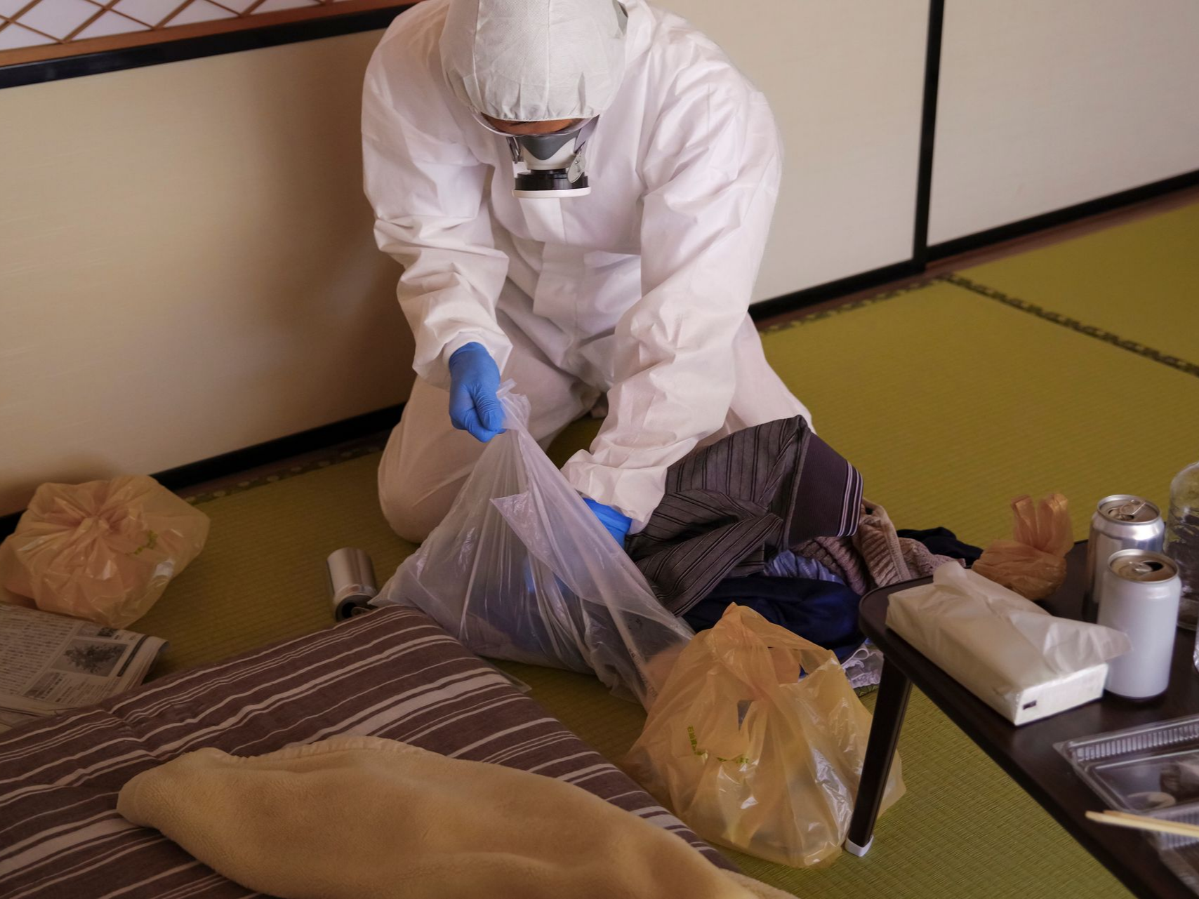 Une personne en combinaison de protection, agenouillée à l'intérieur, met des objets dans un sac en plastique.