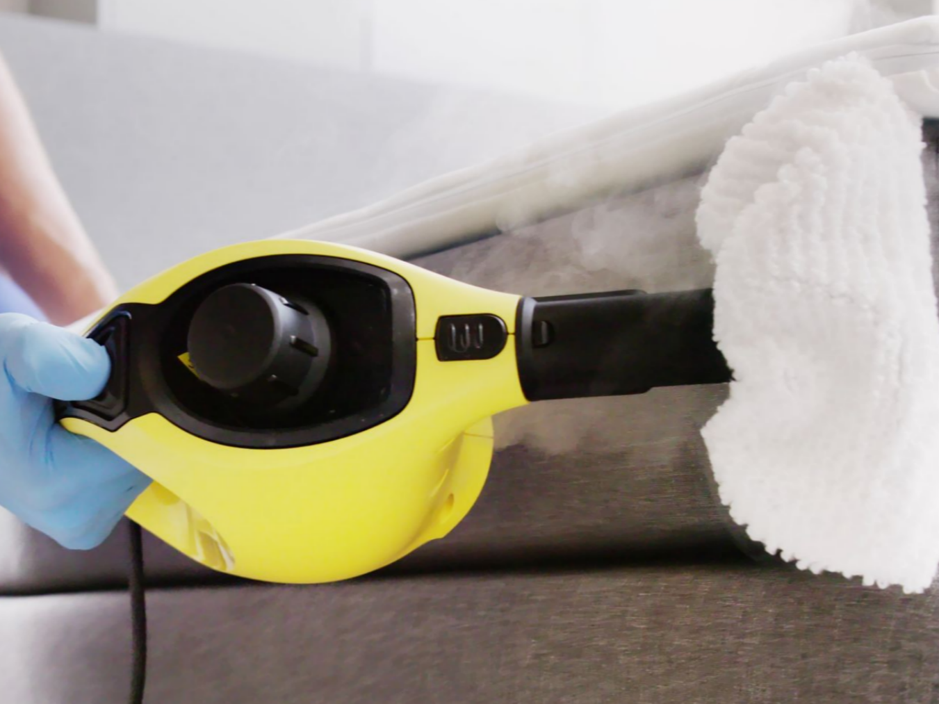 Une personne portant un gant bleu utilise un nettoyeur vapeur sur un canapé.