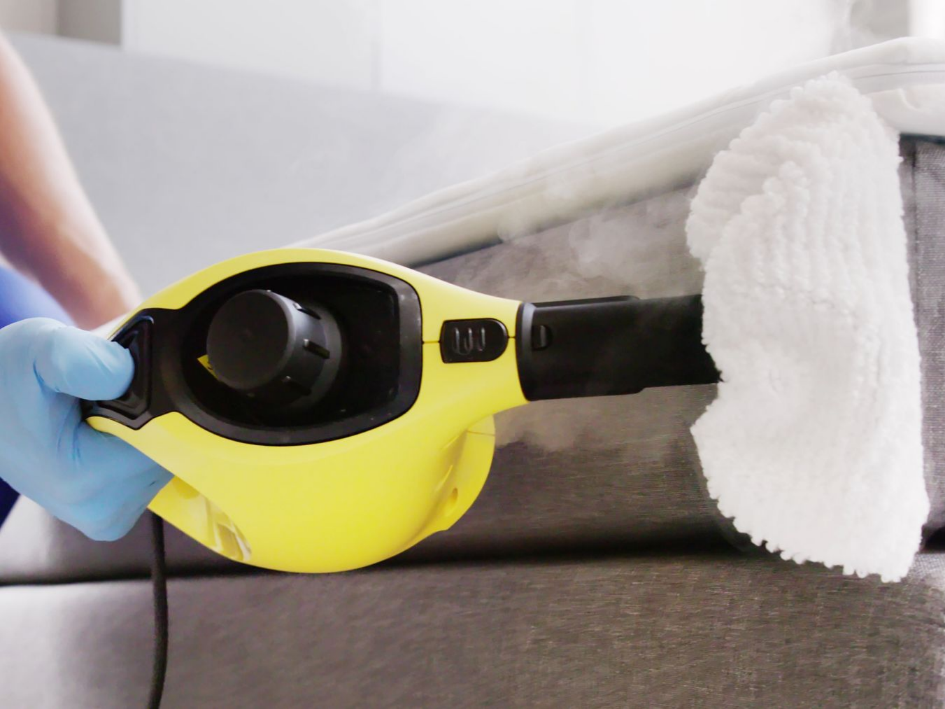 Une personne portant un gant bleu utilise un nettoyeur vapeur pour nettoyer un canapé.