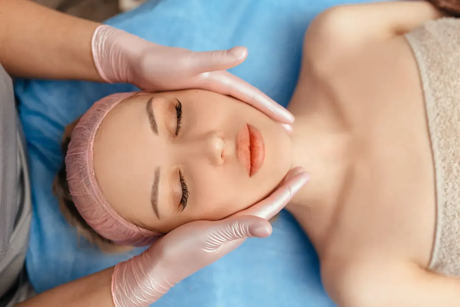 Persona recibiendo un masaje facial en una camilla de spa azul, con una diadema rosa y una toalla puesta.