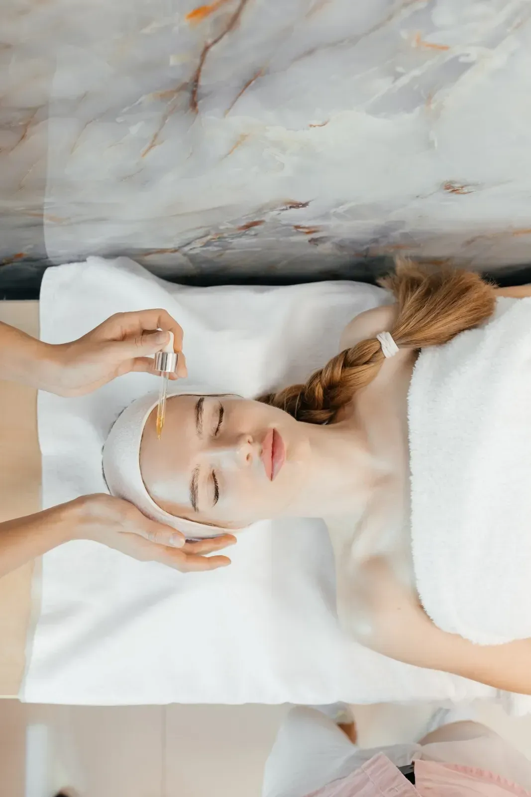 Persona relajándose en un spa con una mascarilla facial, con los ojos cerrados, sobre una toalla blanca y un fondo de mármol.