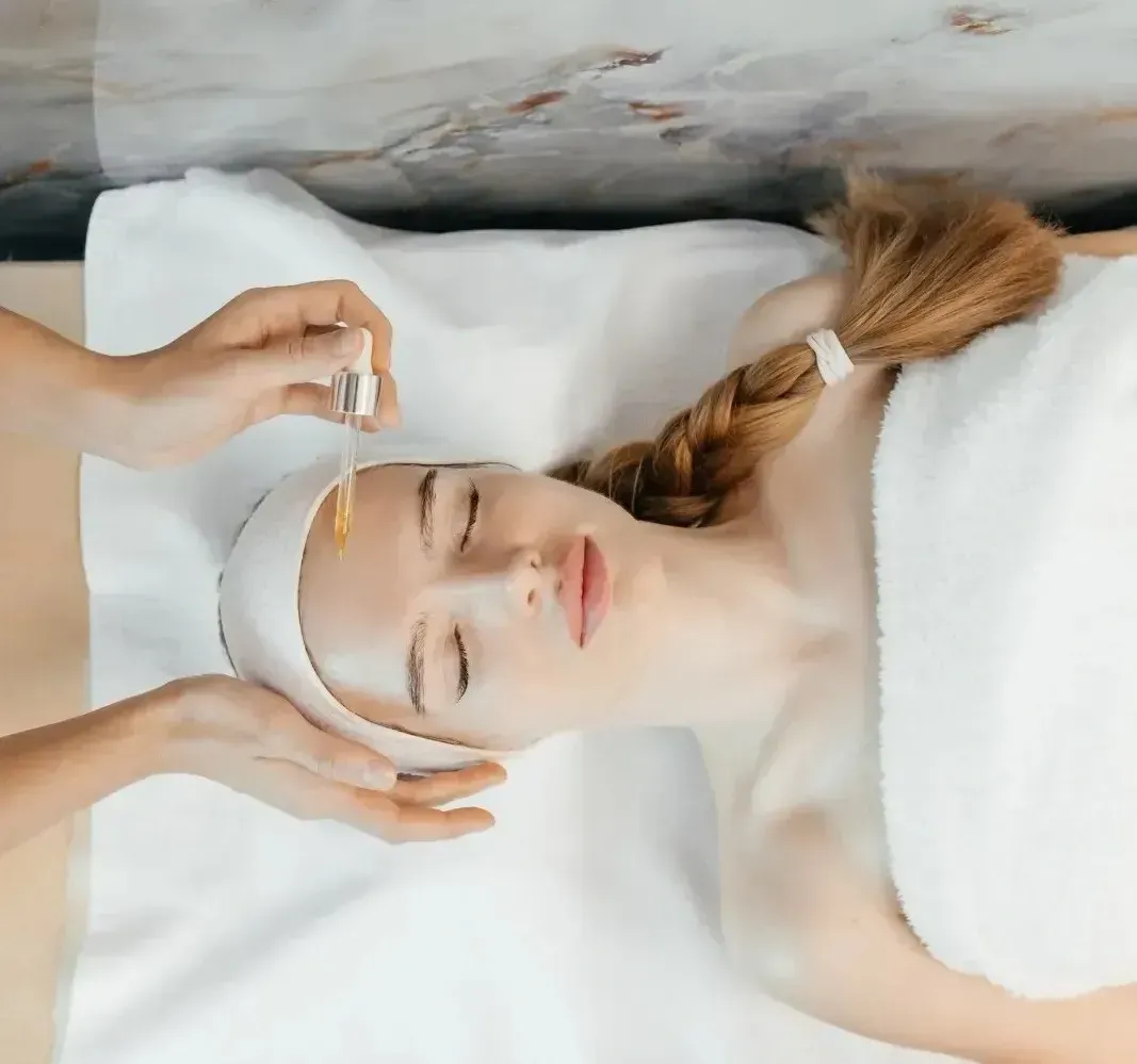 Persona relajándose en un spa con una mascarilla facial, con los ojos cerrados, sobre una toalla blanca y un fondo de mármol.