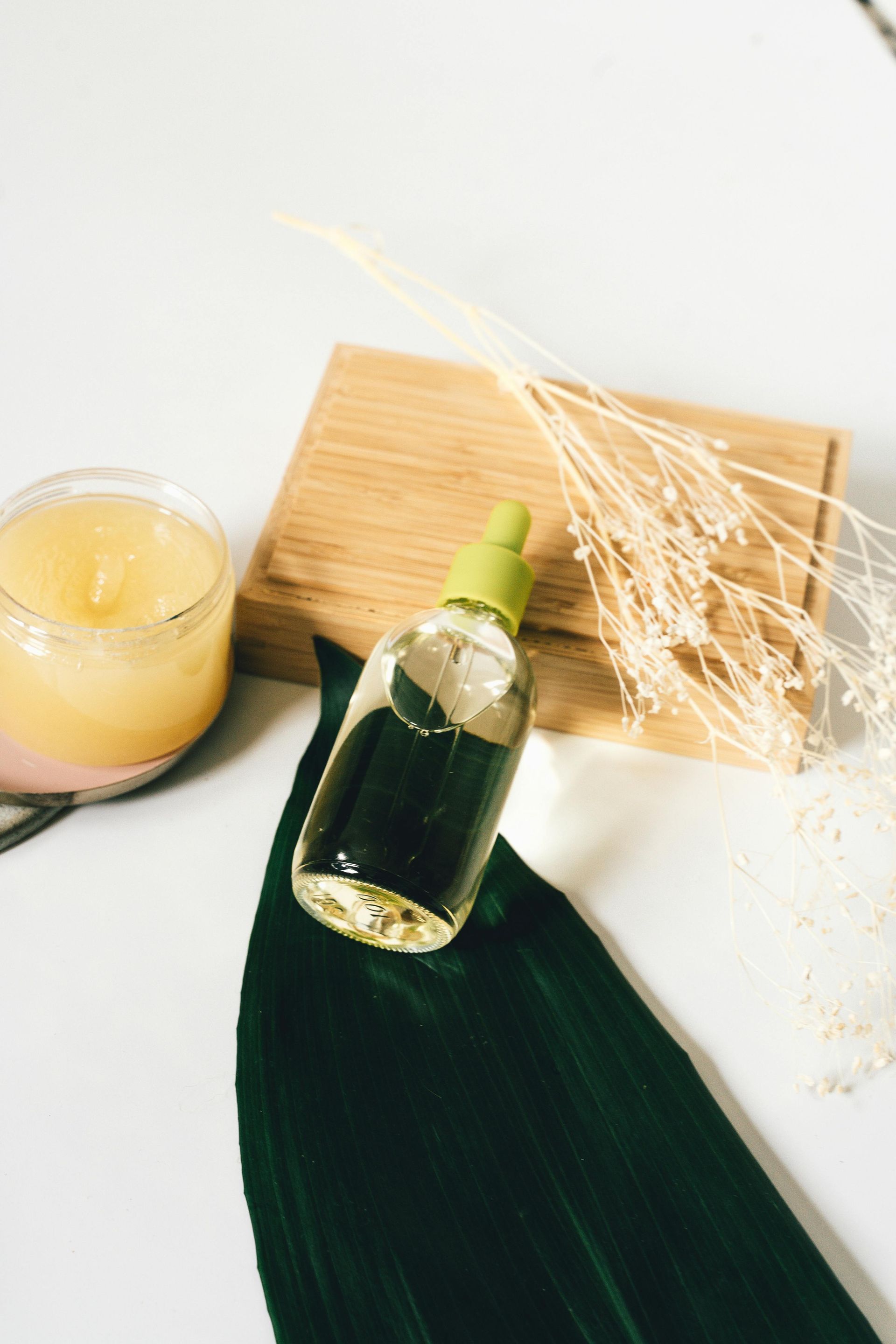 Una botella verde junto a una vela, una tabla de madera y hierba seca sobre una superficie blanca con una hoja oscura.
