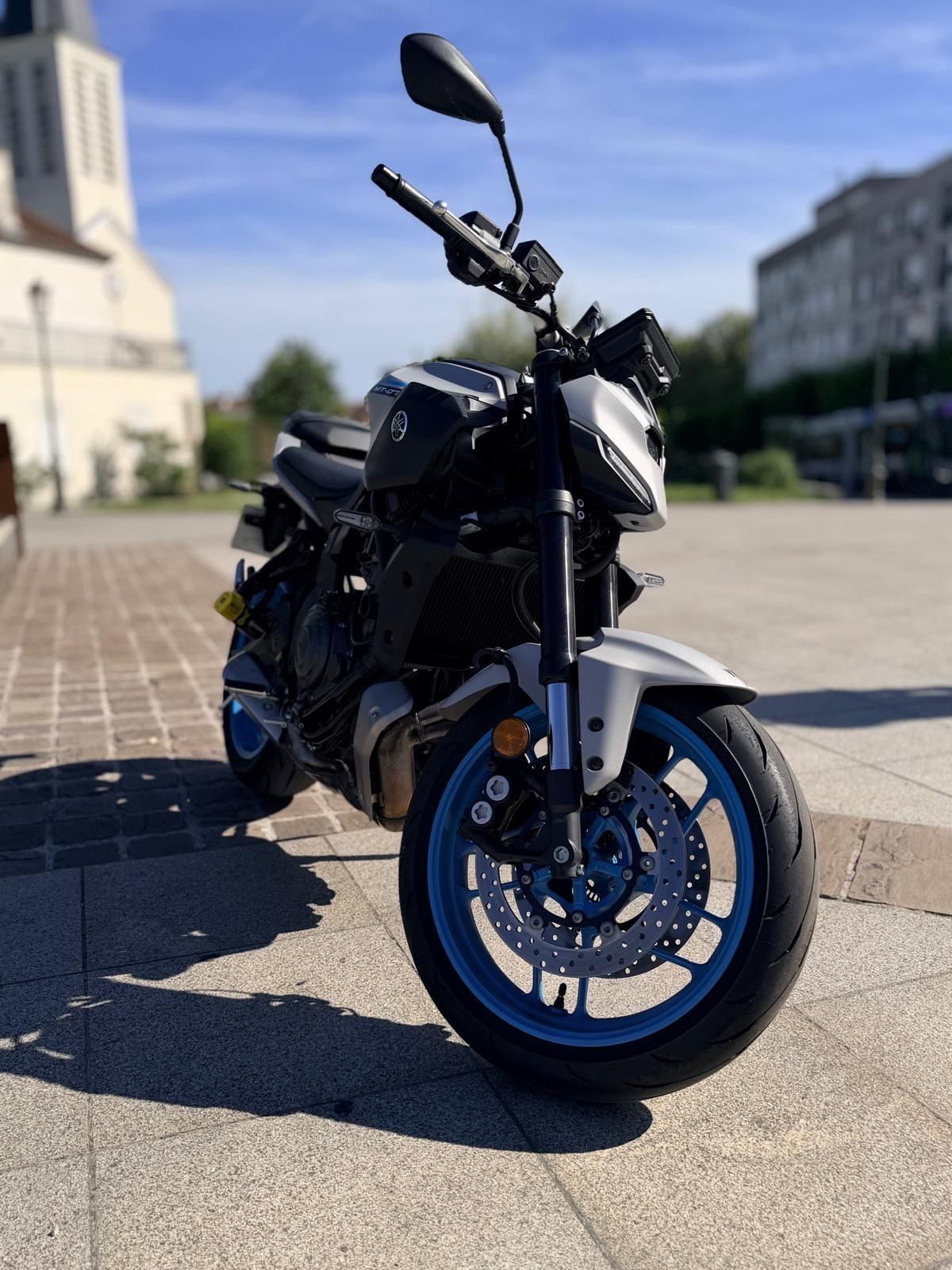 Un scooter noir à pare-brise haut est garé sur le trottoir, près d'une haie et de la route.