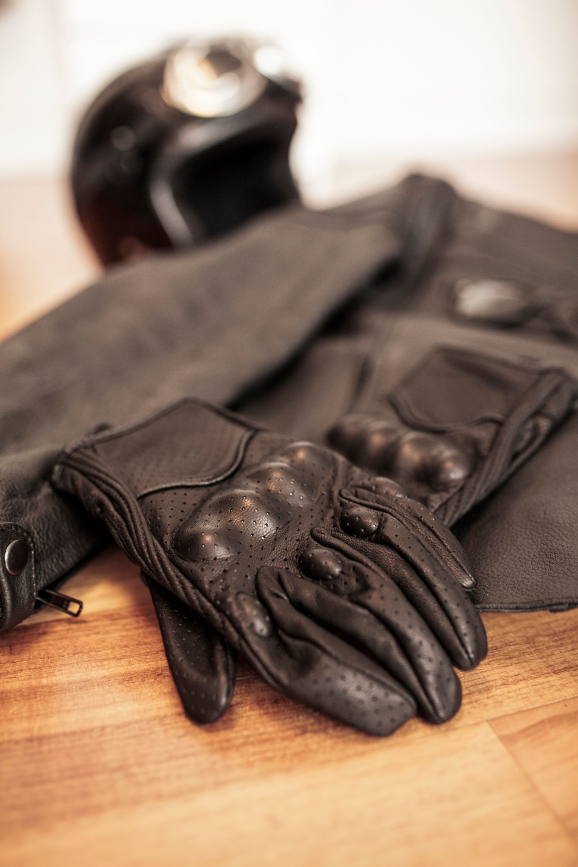 Des gants et un manteau en cuir noir posés sur une surface en bois, avec un casque en arrière-plan.