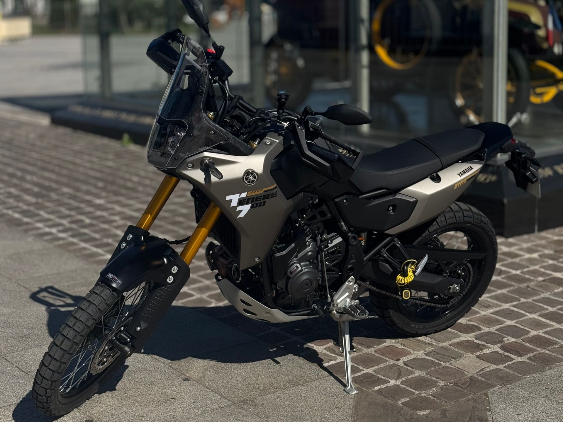 Une moto sport-touring noire est garée sur le trottoir à côté d'une clôture en grillage vert.