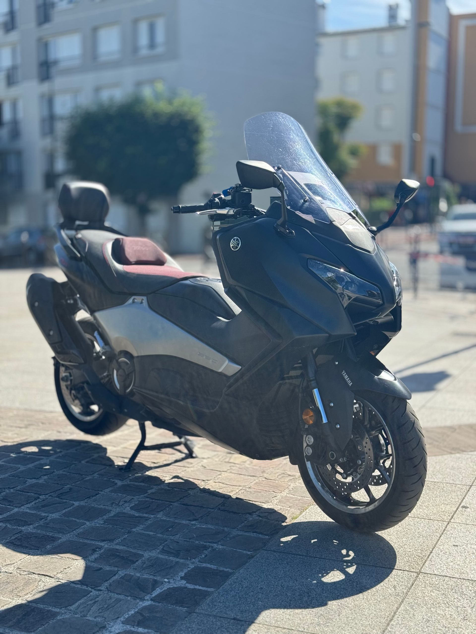 Deux scooters noirs garés dans une allée, à côté d'un mur en bois, l'un d'eux étant équipé d'un pare-brise haut.