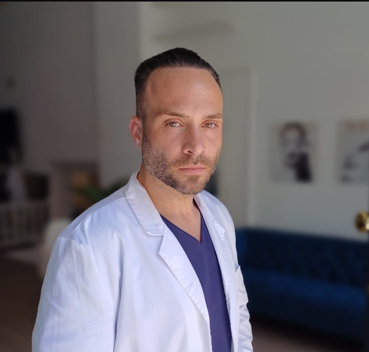 Hombre con bata blanca de laboratorio y uniforme médico azul marino de pie en el interior con expresión neutra, habitación borrosa detrás de él.