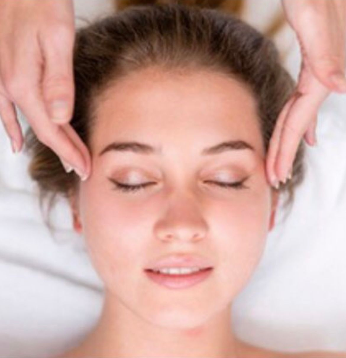 Une femme reçoit un massage du visage, les yeux fermés, les mains sur les tempes.