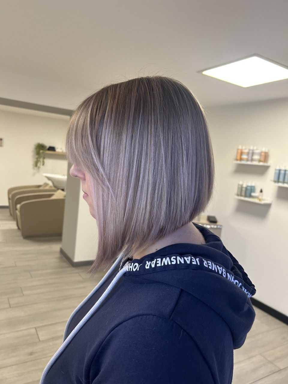 Femme aux cheveux blond cendré dégradés, dans un salon de coiffure, portant un sweat à capuche foncé.