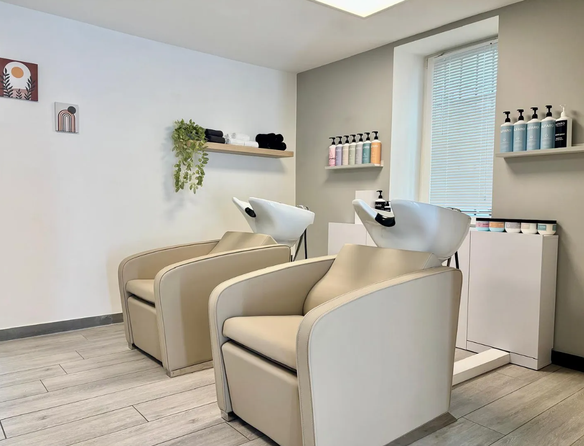 Deux fauteuils de shampoing beiges dans un salon de coiffure avec une fenêtre, des étagères et divers produits capillaires.