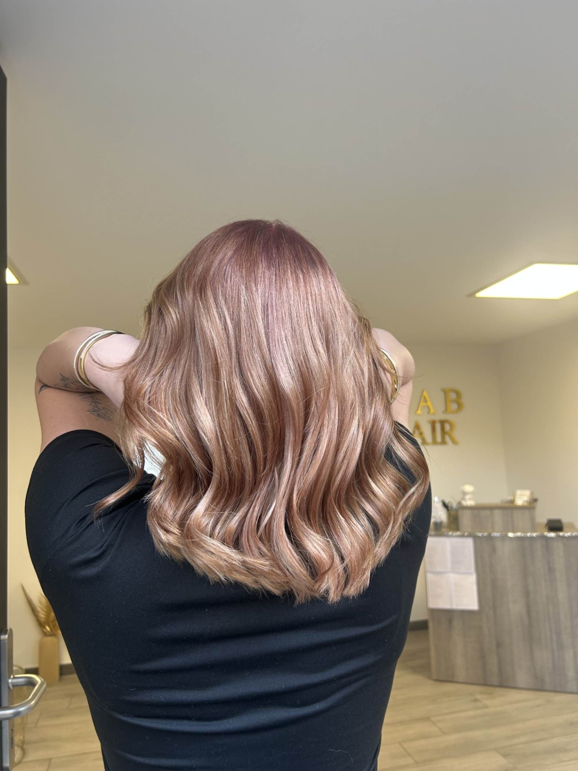 Personne aux cheveux ondulés, blond rosé. Haut foncé, mains près des cheveux. Intérieur d'un salon de coiffure.