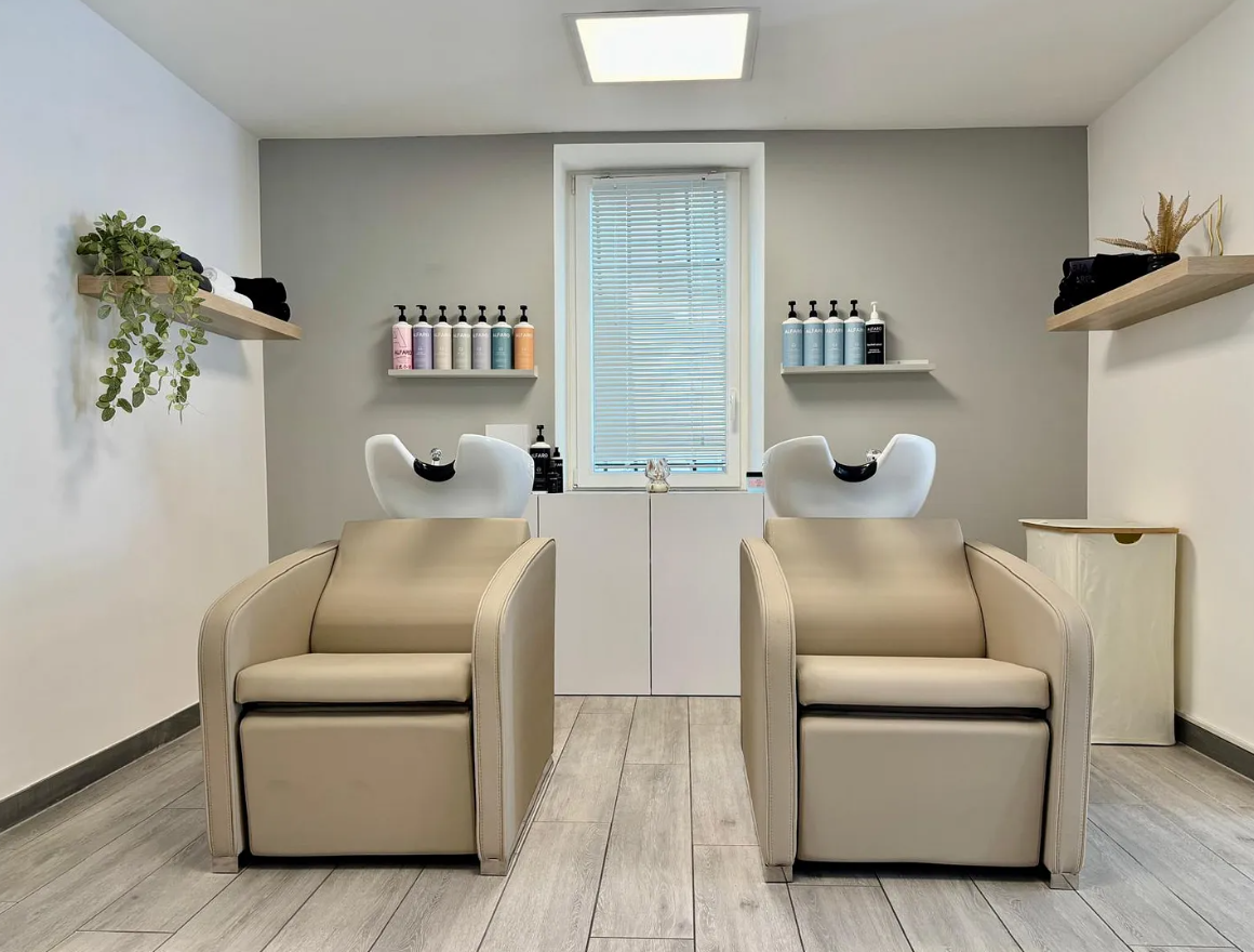 Deux fauteuils de shampoing beiges avec lavabos, des murs gris, des étagères avec des produits et une fenêtre.