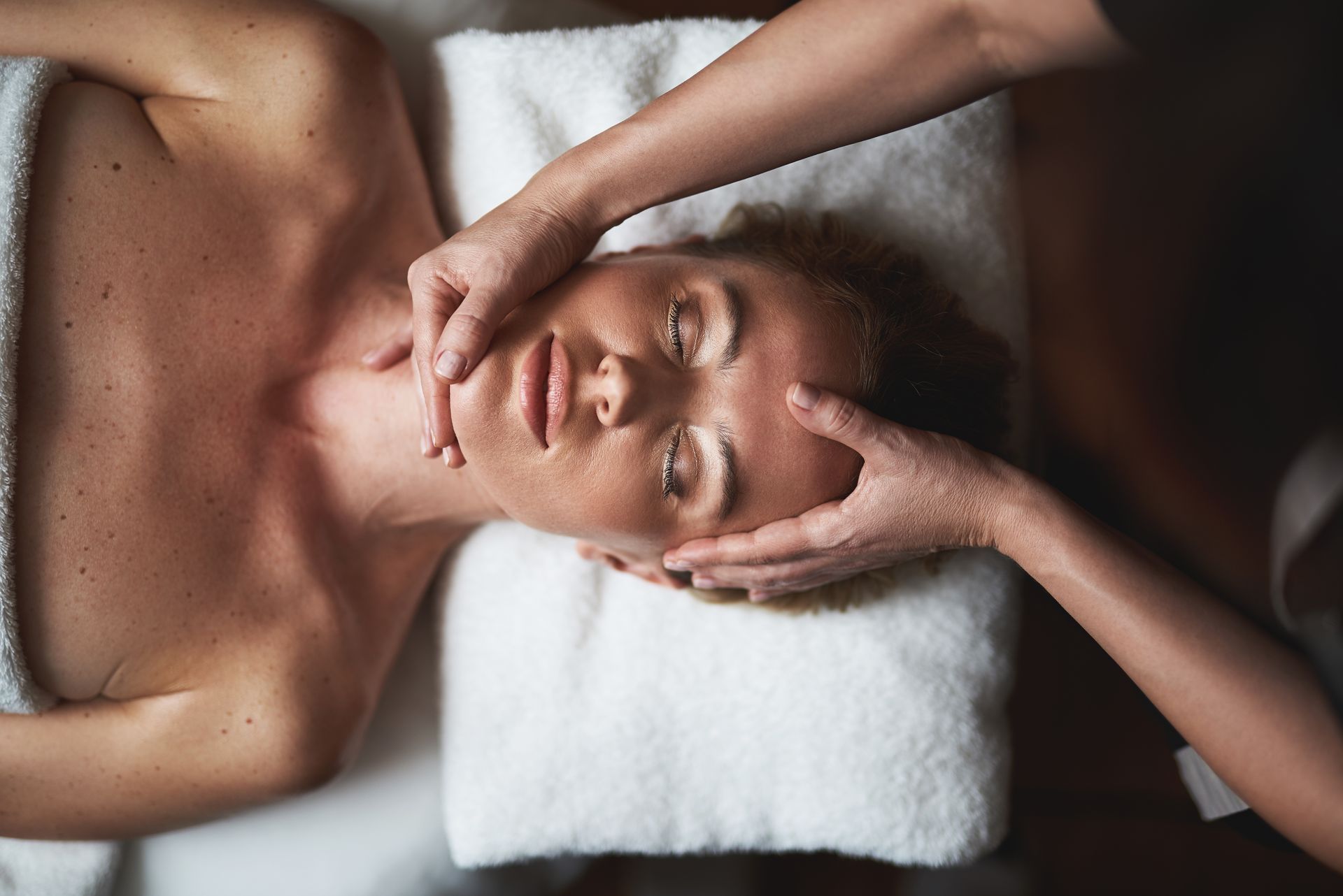 Une personne reçoit un massage du visage sur une table de massage, une serviette blanche sous la tête.