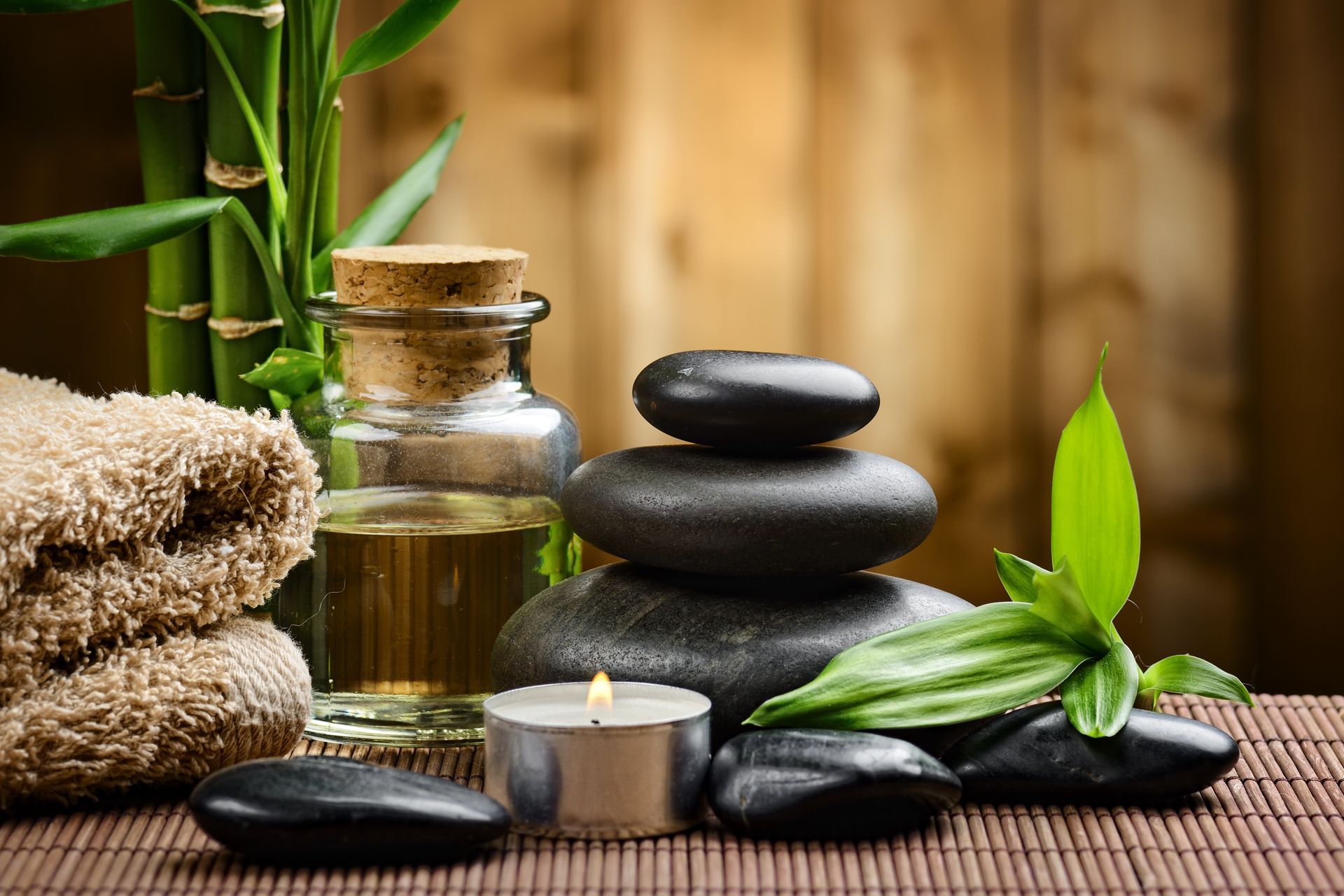 Aménagement spa avec bambou, pierres empilées, bougie, flacon d'huile, serviettes et feuilles vertes sur un tapis tissé.