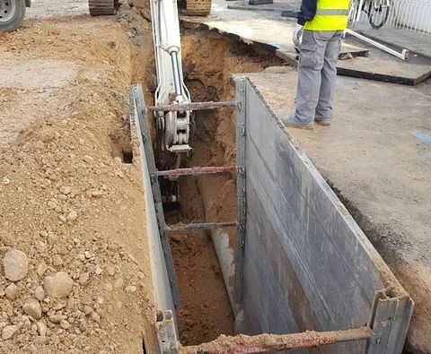Un hombre con un chaleco amarillo está parado en una zanja al lado de una excavadora.