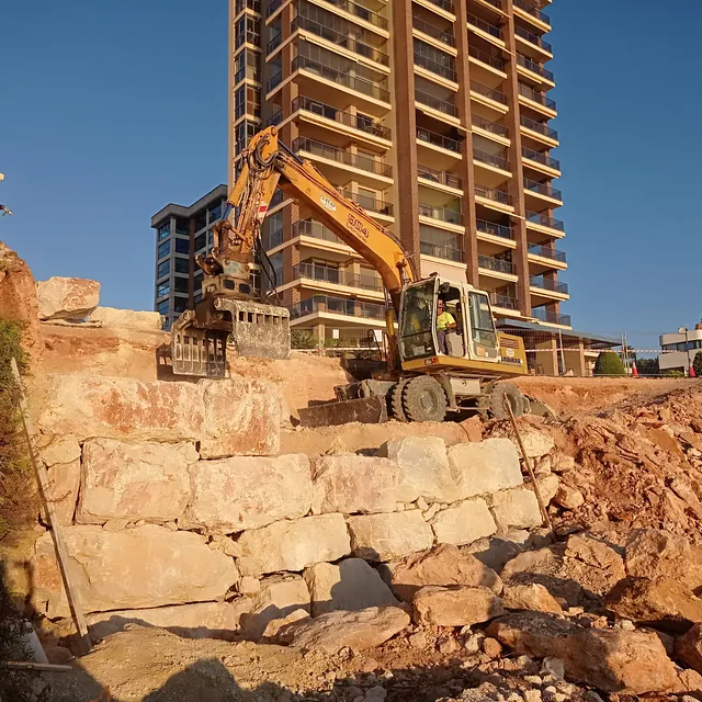 Una excavadora está trabajando en un muro de piedra frente a un edificio alto.