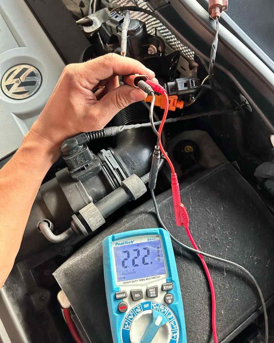Une personne utilise un multimètre pour tester un composant automobile dans le compartiment moteur. L'appareil affiche 222.