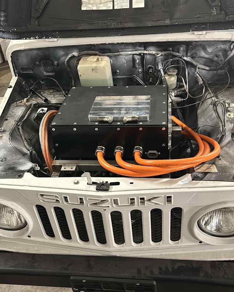 Suzuki Samurai blanche, capot ouvert, laissant apparaître des composants de véhicule électrique dans le compartiment moteur.