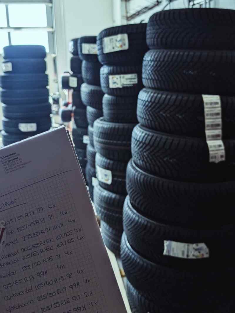 Dans un entrepôt, des piles de pneus noirs étiquetés ; au premier plan, une personne tient des documents.