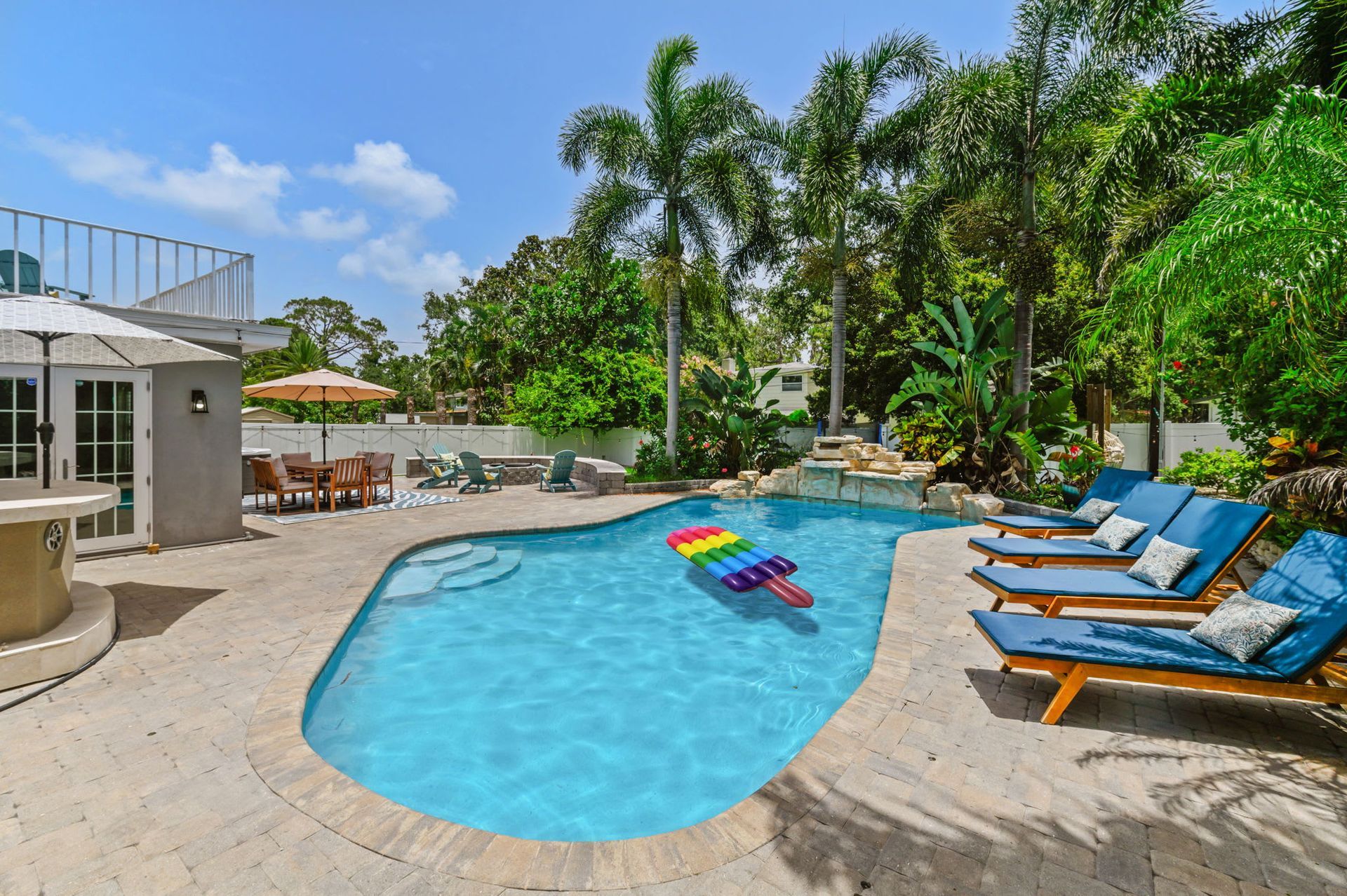 Heated pool with waterfall, sun loungers, and lush tropical vibes—your private backyard escape. Luxury backyard with heated swimming pool, waterfall feature, palm trees, and blue lounge chairs in a tropical setting.