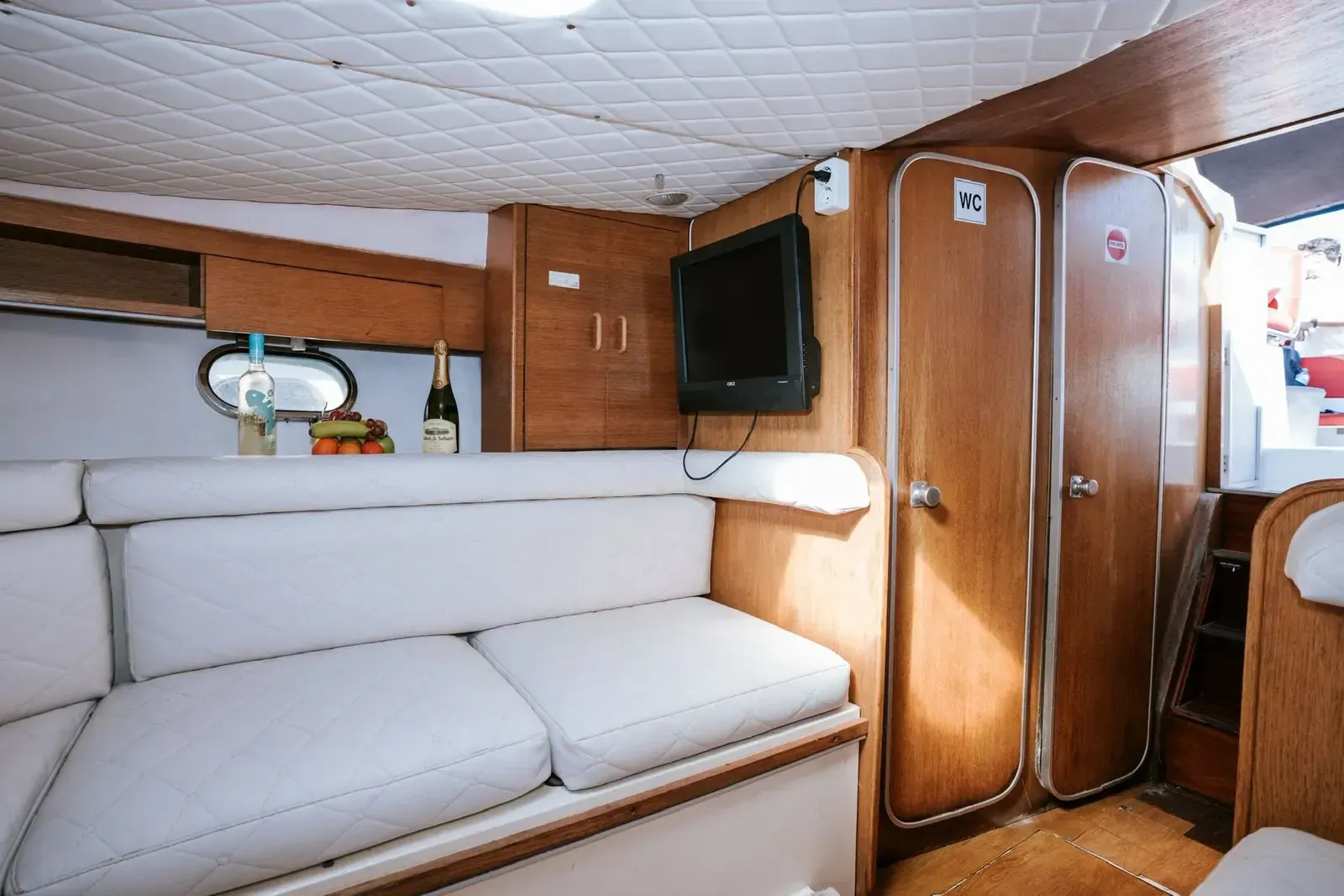 Interior de una cabina de barco con asientos blancos, paneles de madera y un televisor montado.